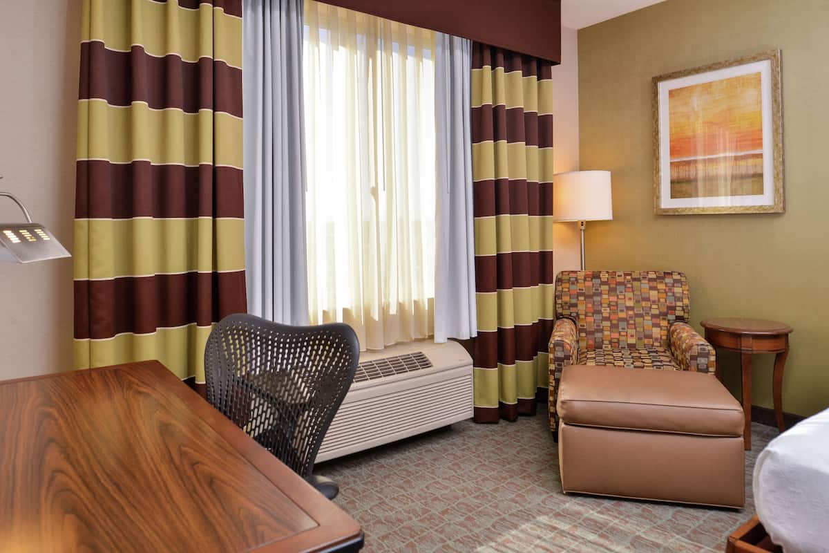 A hotel room with striped curtains, a patterned armchair, a wooden table, a small round table, and a wall-mounted lamp.