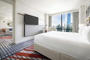Quarto de hotel moderno com uma cama grande, televisão de tela plana na parede, escrivaninha com cadeira e vista para o horizonte da cidade através de grandes janelas.