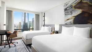 Ein Hotelzimmer mit zwei Betten, einem Schreibtisch und einem Stuhl. Durch große Fenster ist die Stadtlandschaft zu sehen.