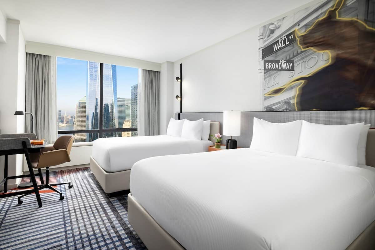 Ein Hotelzimmer mit zwei Betten, einem Schreibtisch und einem Stuhl. Durch große Fenster ist die Stadtlandschaft zu sehen.
