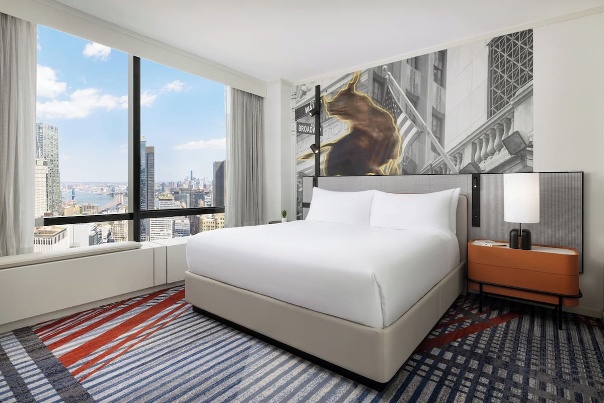 Ein Hotelzimmer mit einem großen Bett, einem Nachttisch, einer Lampe und Blick auf die Stadt durch das Fenster.