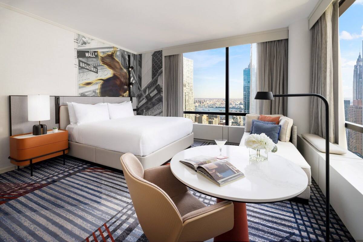 Ein Hotelzimmer mit einem großen Bett, einem runden Tisch mit einer Zeitschrift, einem Sessel, einem Nachttisch und Blick auf die Stadt.