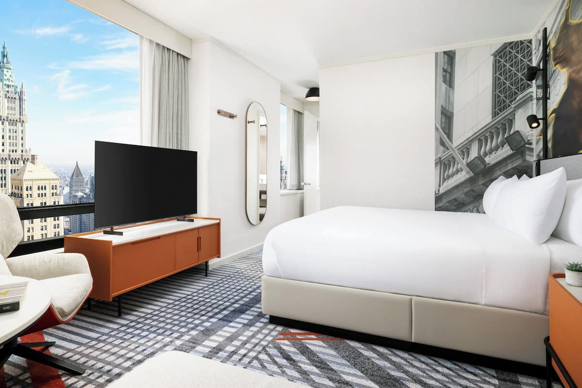 Ein Hotelzimmer mit einem großen Bett, einem Fernseher auf einer Holzkonsole, Blick auf die Stadt und einem gemusterten Teppich.
