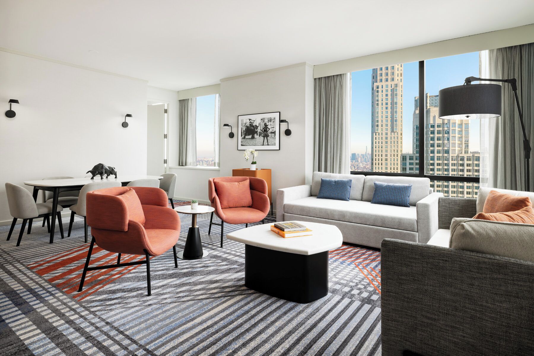 A modern hotel room with a white sofa, two armchairs, a dining table with chairs, a coffee table, and large windows offering a city view.