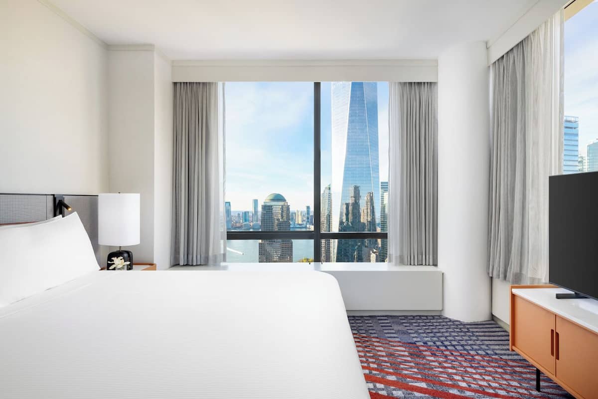 Ein Hotelzimmer mit einem großen Bett, einem Fernseher, einem Schreibtisch und Blick auf eine Skyline durch raumhohe Fenster.