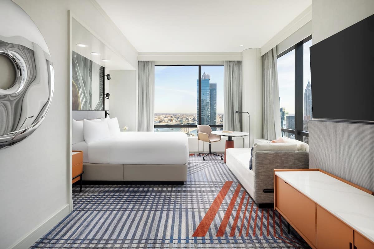 Ein Hotelzimmer mit einem großen Bett, einem Schreibtisch, einem Sessel und einer Couch. Große Fenster bieten Blick auf die Stadt.