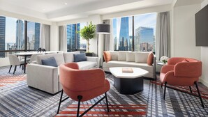Ein modernes Hotelzimmer mit großem Fenster und Blick auf die Stadt, einer Sofagarnitur, einem Couchtisch und einem gemusterten Teppich.