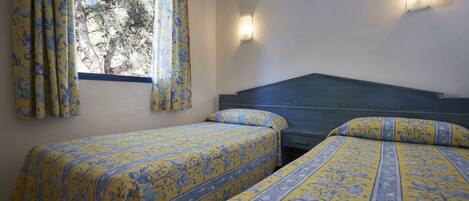 A room with two beds, each with a floral-patterned bedspread, a window with curtains, and a wall-mounted light fixture.