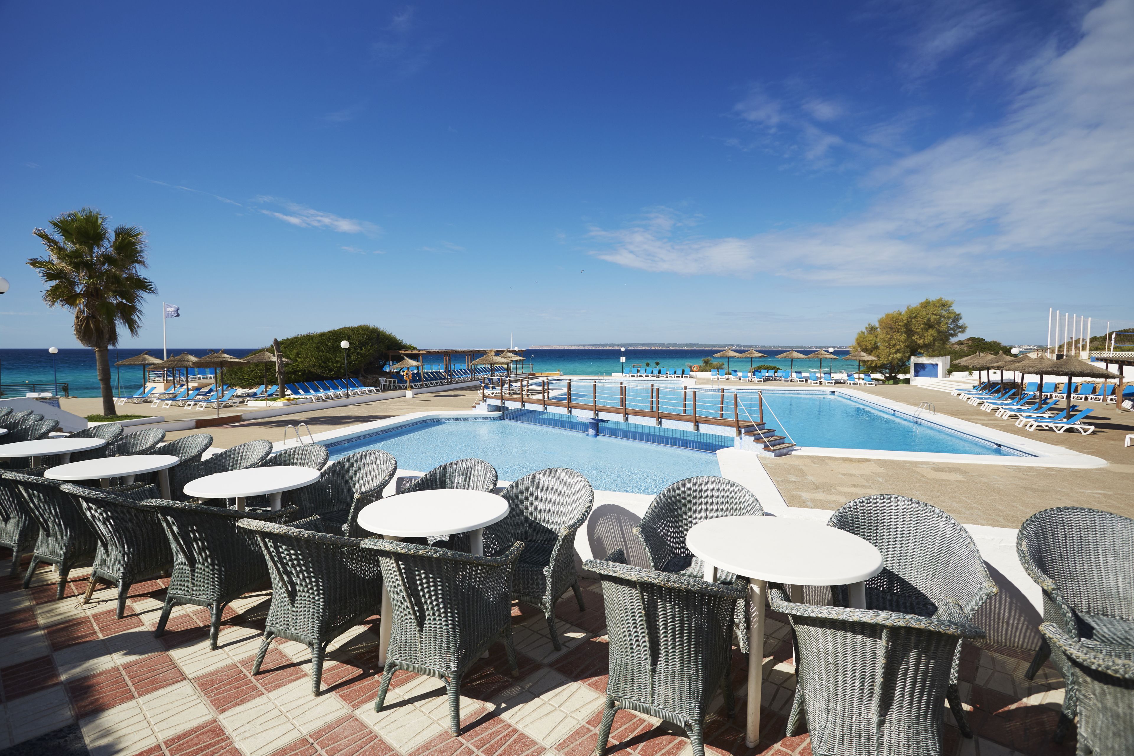 A poolside area with wicker chairs and tables, a swimming pool, and a view of the sea.