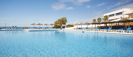 A large swimming pool with blue water, surrounded by lounge chairs and umbrellas, and a building in the background.