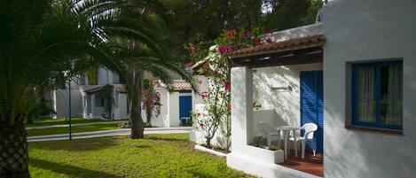 Un edificio blanco con contraventanas azules, un pequeño patio con una mesa y sillas, y una palmera en primer plano.