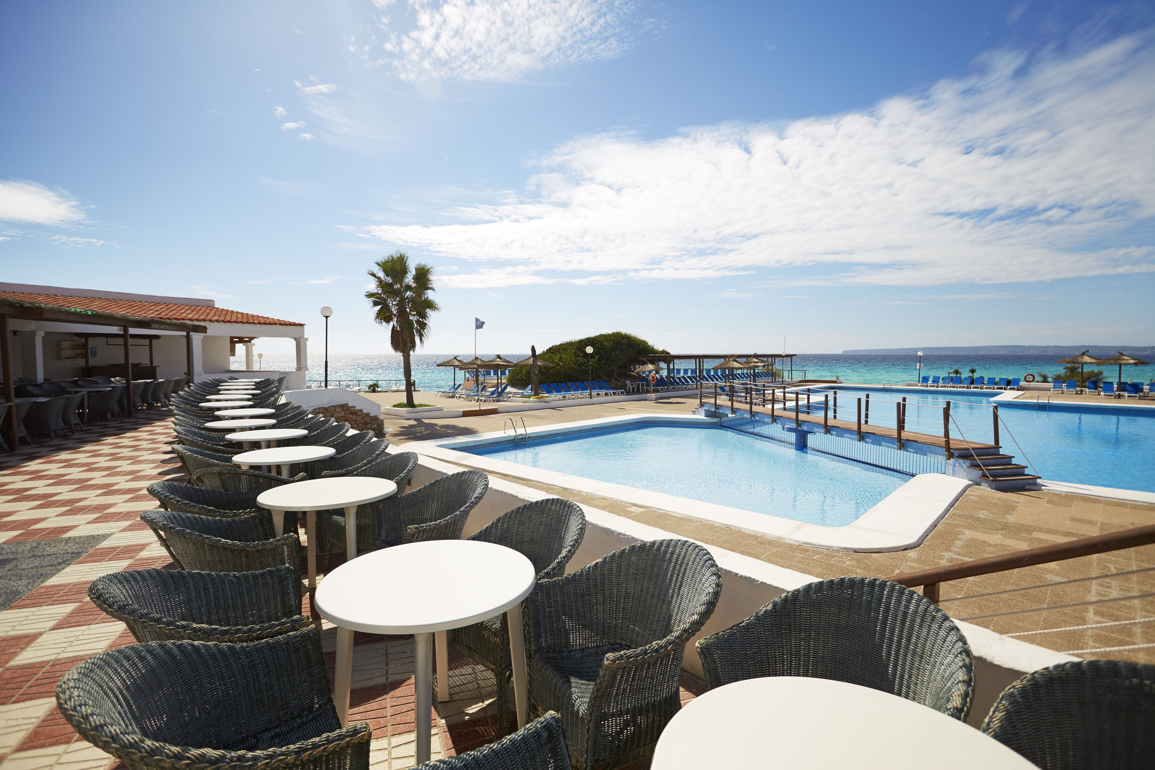 A poolside area with wicker chairs and tables, a pool, and a view of the sea.
