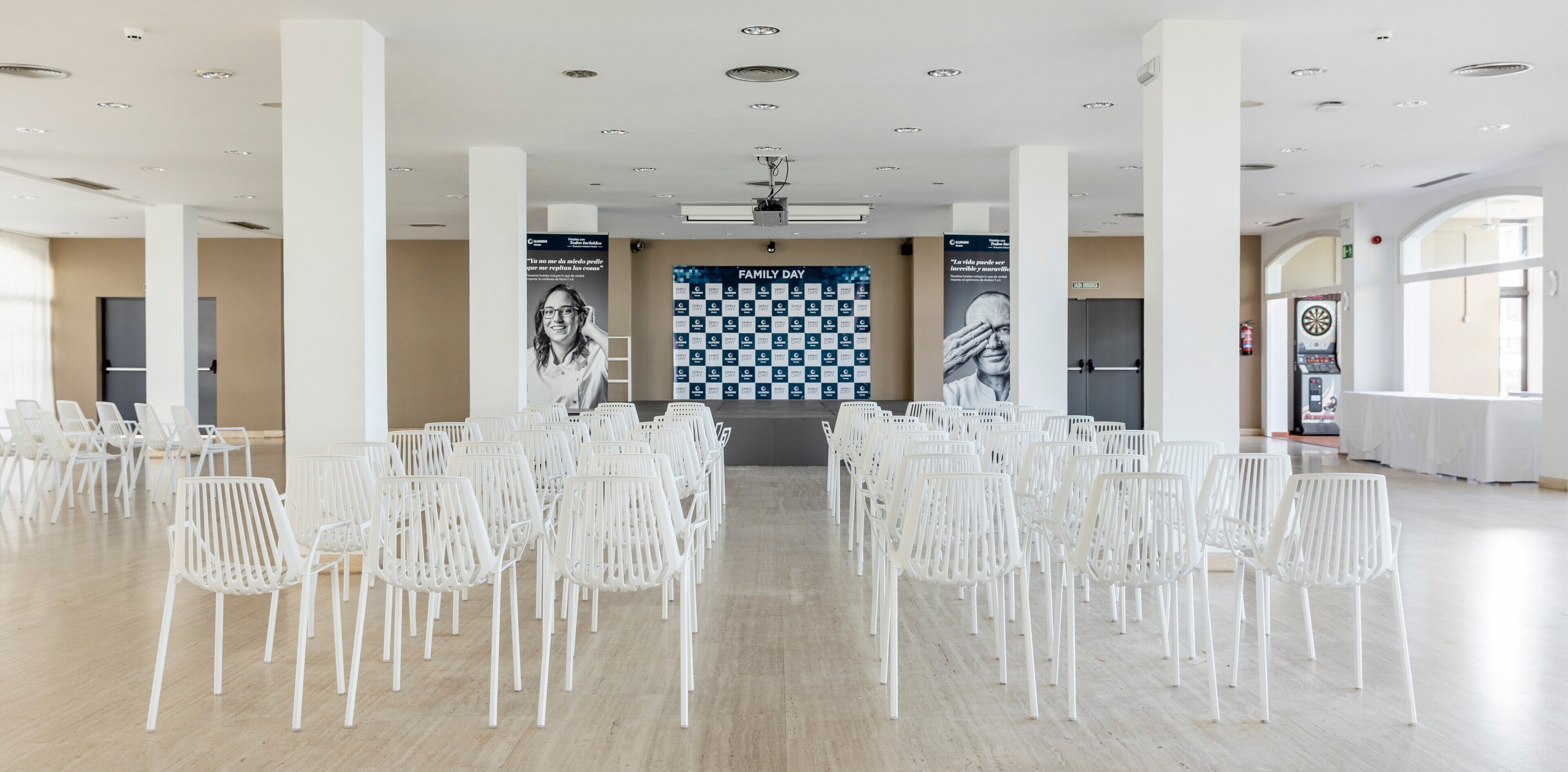 Un salón amplio con filas de sillas blancas, un gran muro a cuadros y un dardo.