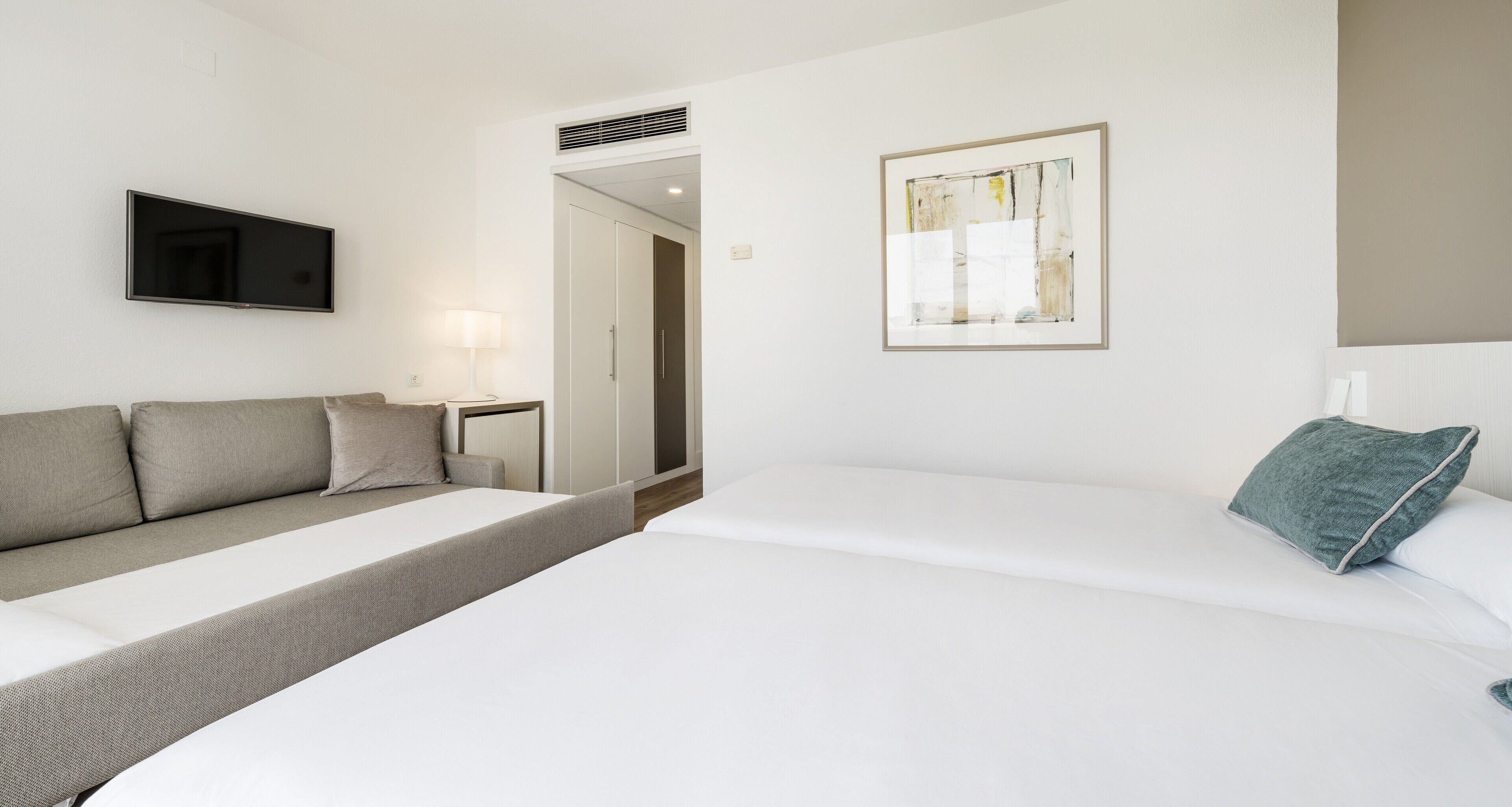A modern hotel room with a sofa, a bed, a flat-screen TV, a framed abstract painting, and a nightstand with a lamp.