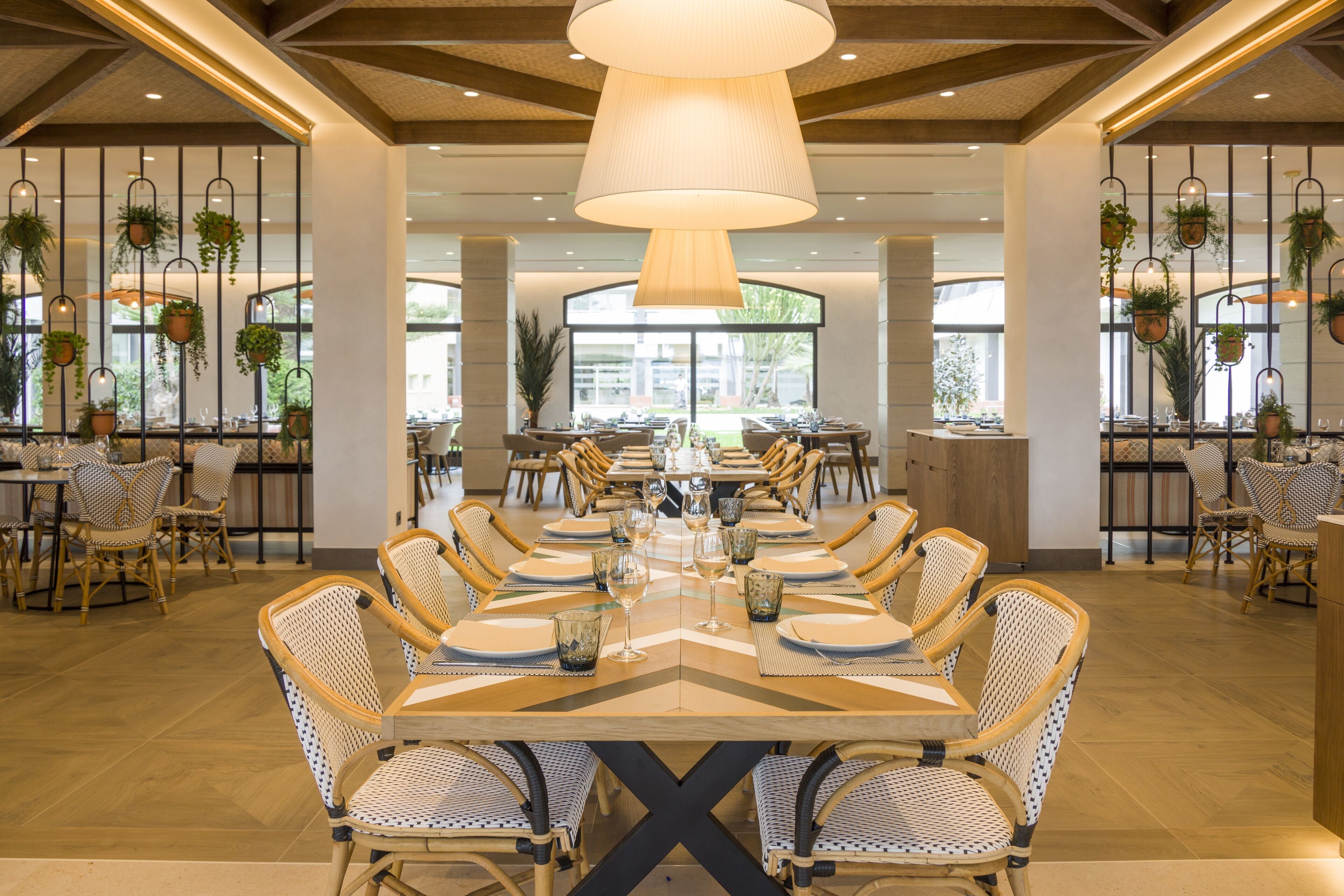 A spacious dining area with a long table set for a meal, surrounded by wicker chairs, under a large hanging light fixture.