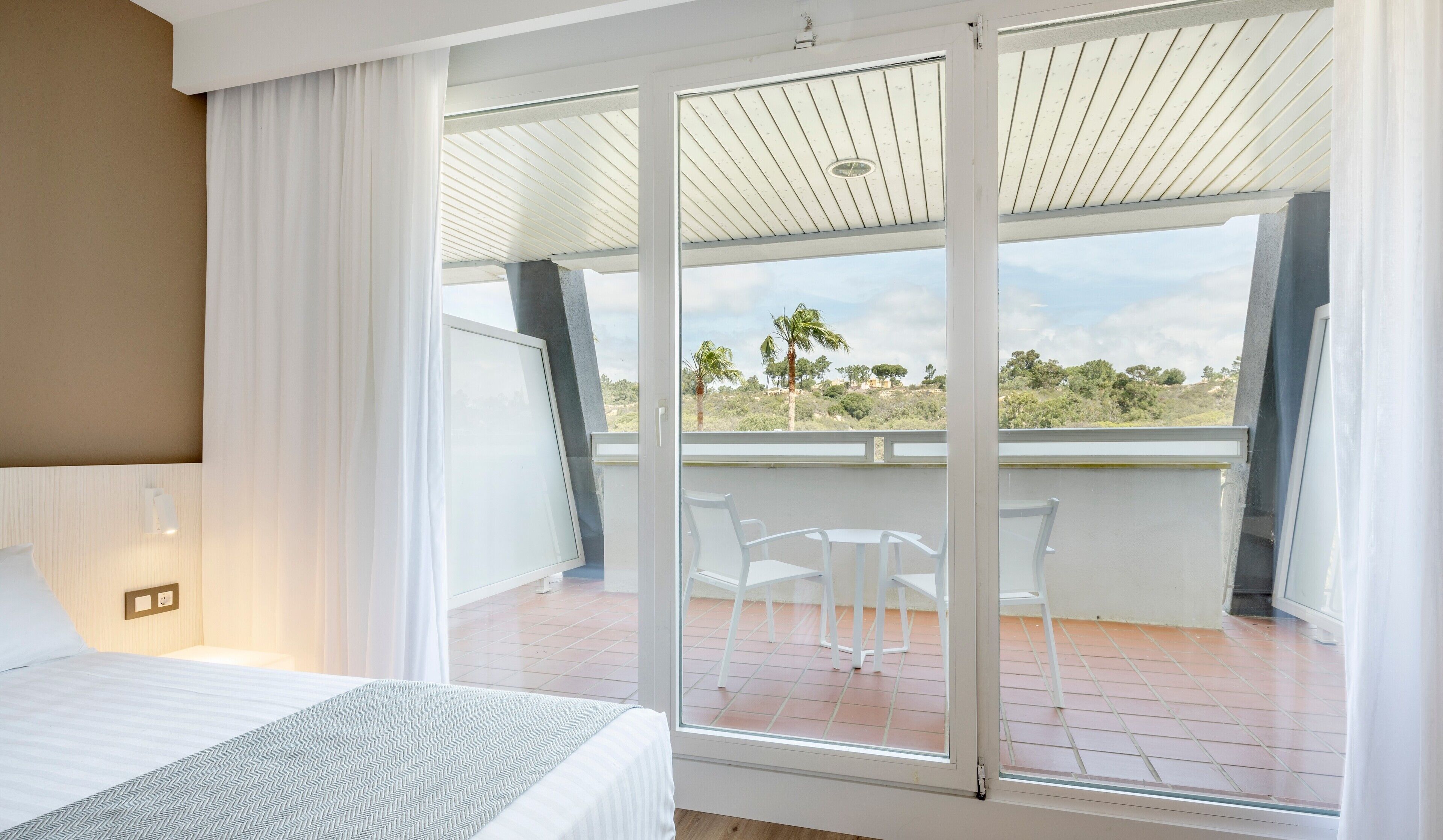 Quarto com uma grande porta de vidro deslizante que dá para uma varanda com cadeiras brancas e vista para palmeiras e colinas.