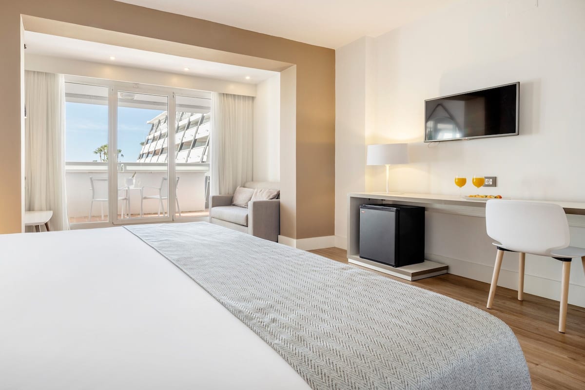 Ein modernes Hotelzimmer mit einem großen Bett, einem Schreibtisch mit Computer, einem Fernseher und einem Balkon mit Blick auf Gebäude.