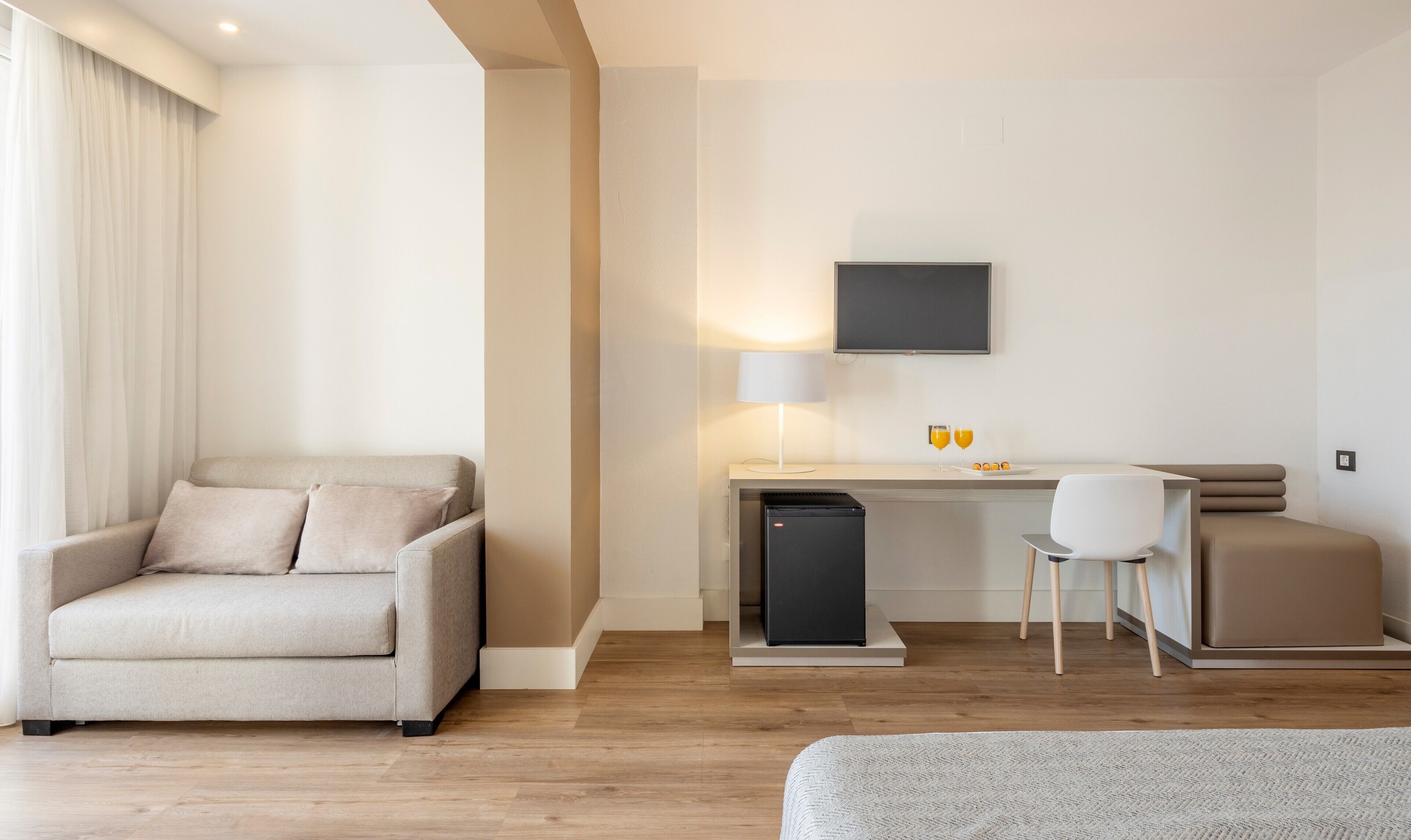 Quarto de hotel moderno com sofá, escrivaninha com computador, televisão e mini-frigorífico.