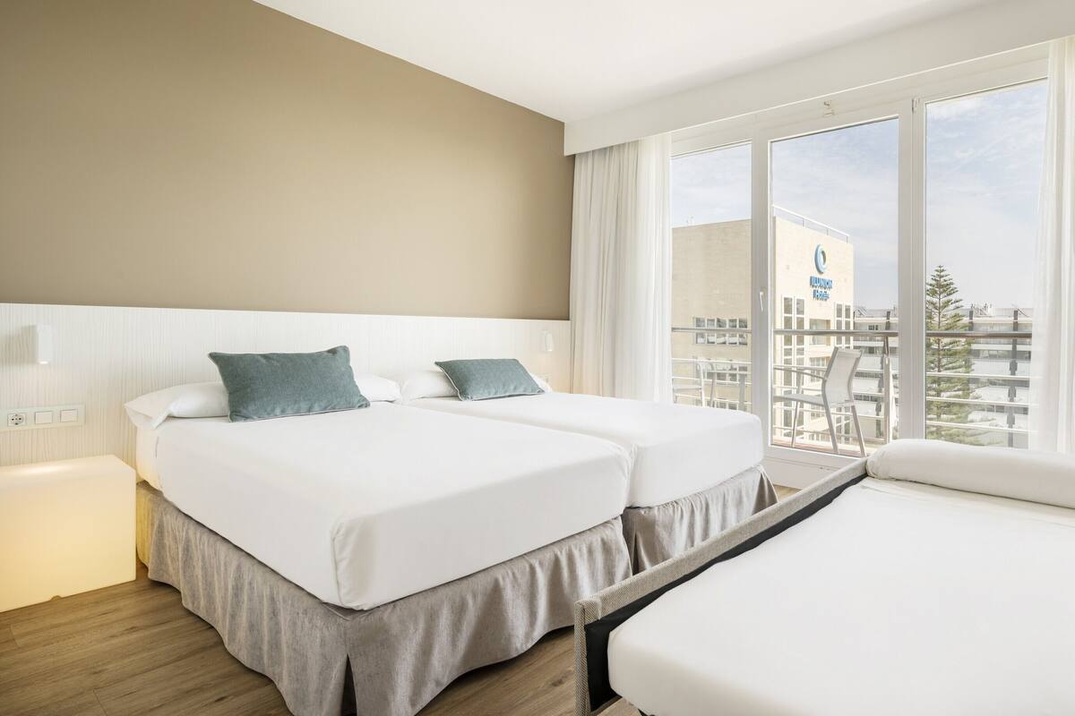 Ein Hotelzimmer mit zwei Betten, einem Sofa und einem großen Fenster mit Blick auf ein Gebäude und Bäume.