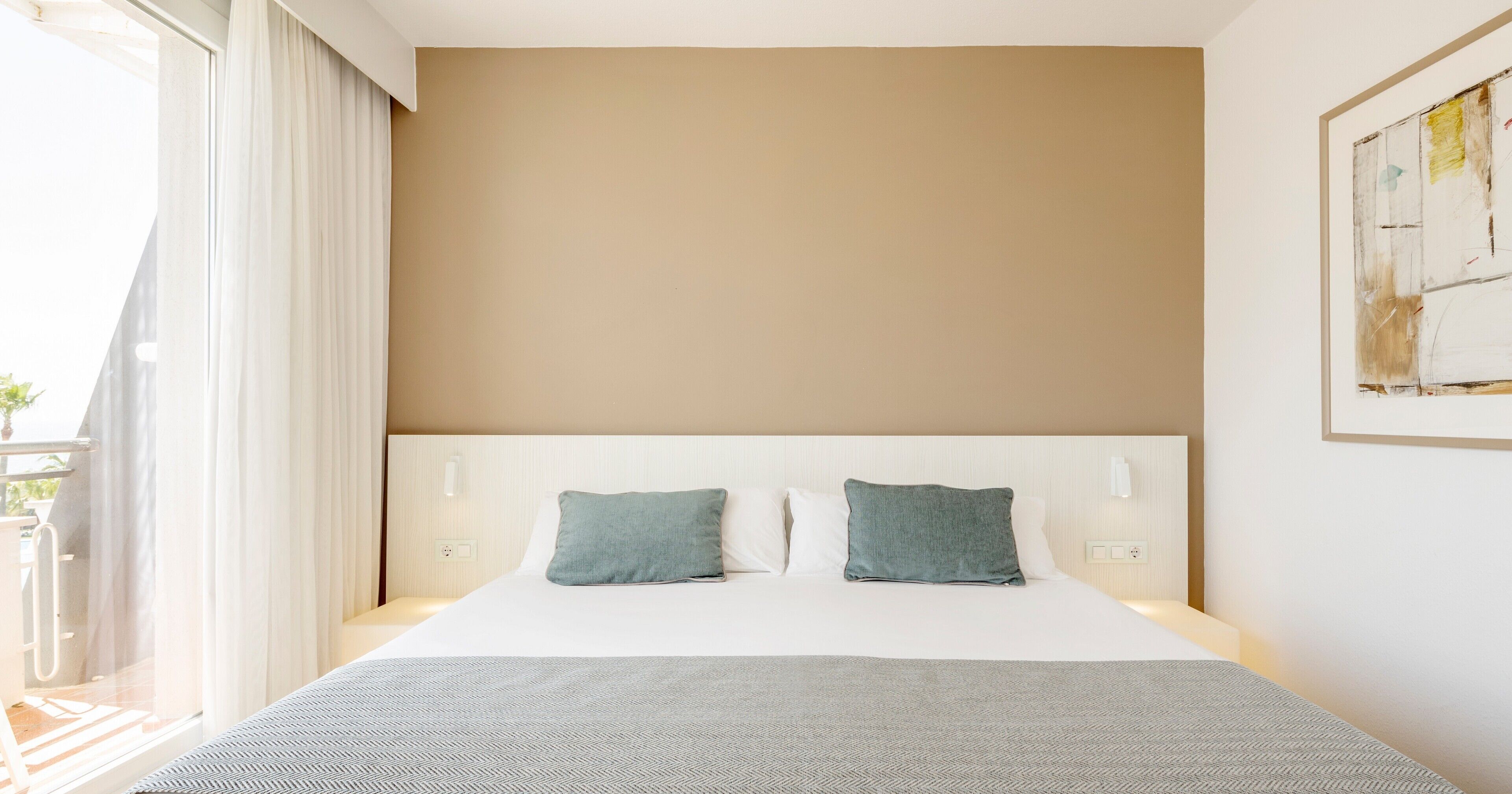 A hotel room with a large bed, two pillows, a gray blanket, and a view of the outdoors.