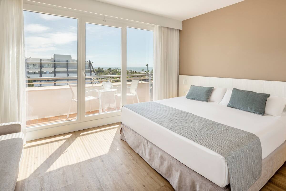 Ein Hotelzimmer mit einem großen Bett, einem Balkon mit weißen Stühlen und Blick auf die Stadt.