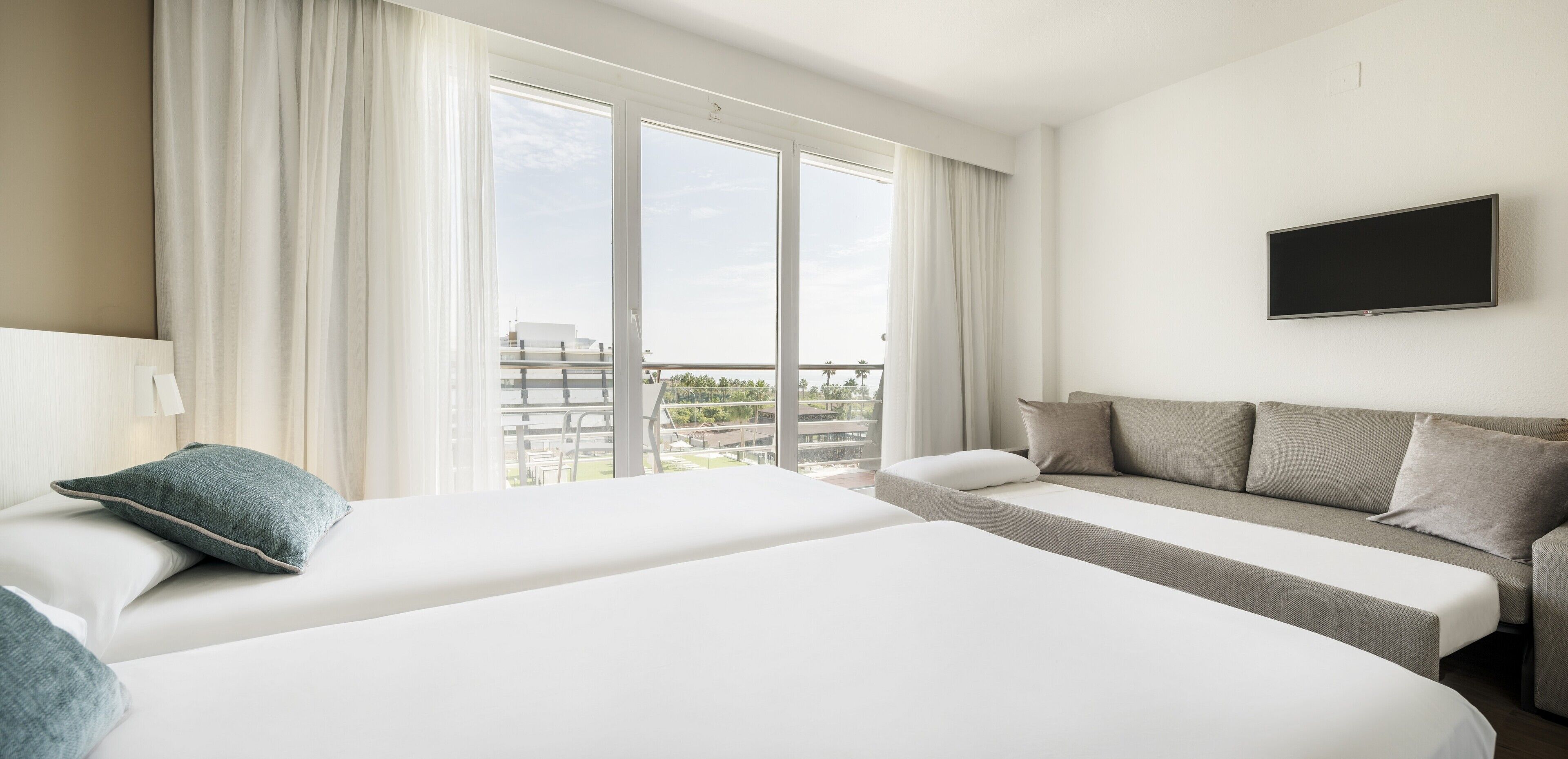 A hotel room with two beds, a sofa, a TV, and large windows with curtains.