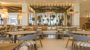 A modern restaurant interior with high ceilings, hanging plants, and a mix of wooden and upholstered furniture. - Hotel ILUNION Islantilla (Islantilla)