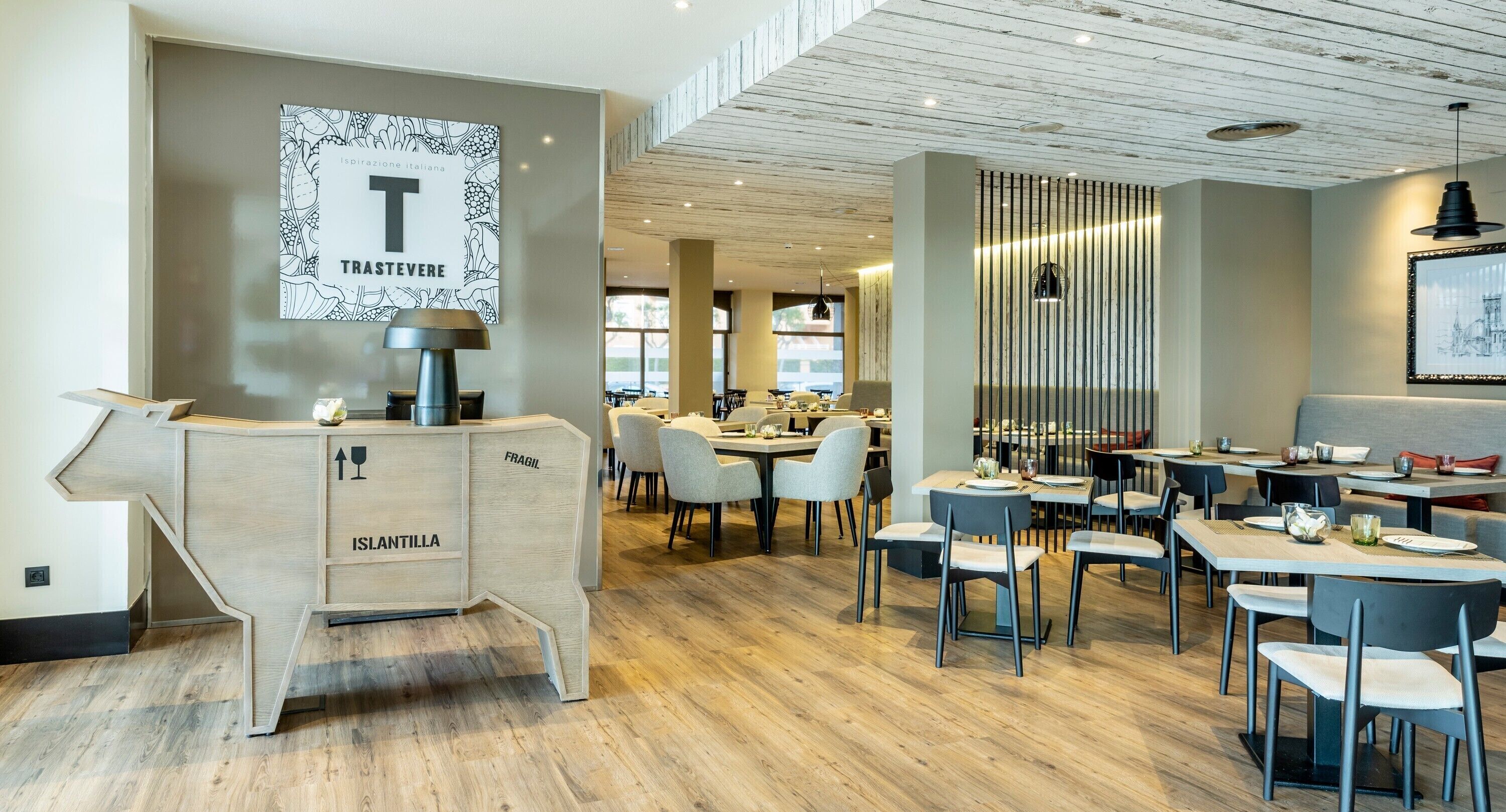 A modern restaurant interior with a wooden counter labeled "FRAGIL" and "ISLANDTILLA," a large framed artwork on the wall, and a seating area with tables and chairs.