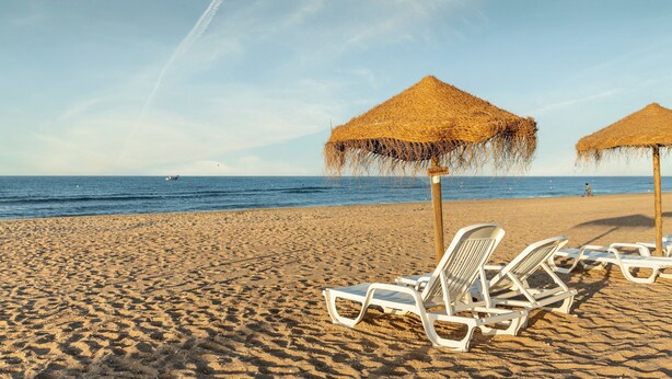 A beach with sun loungers and thatched umbrellas, a clear sky, and a boat in the distance. - Hotel ILUNION Islantilla (Islantilla)