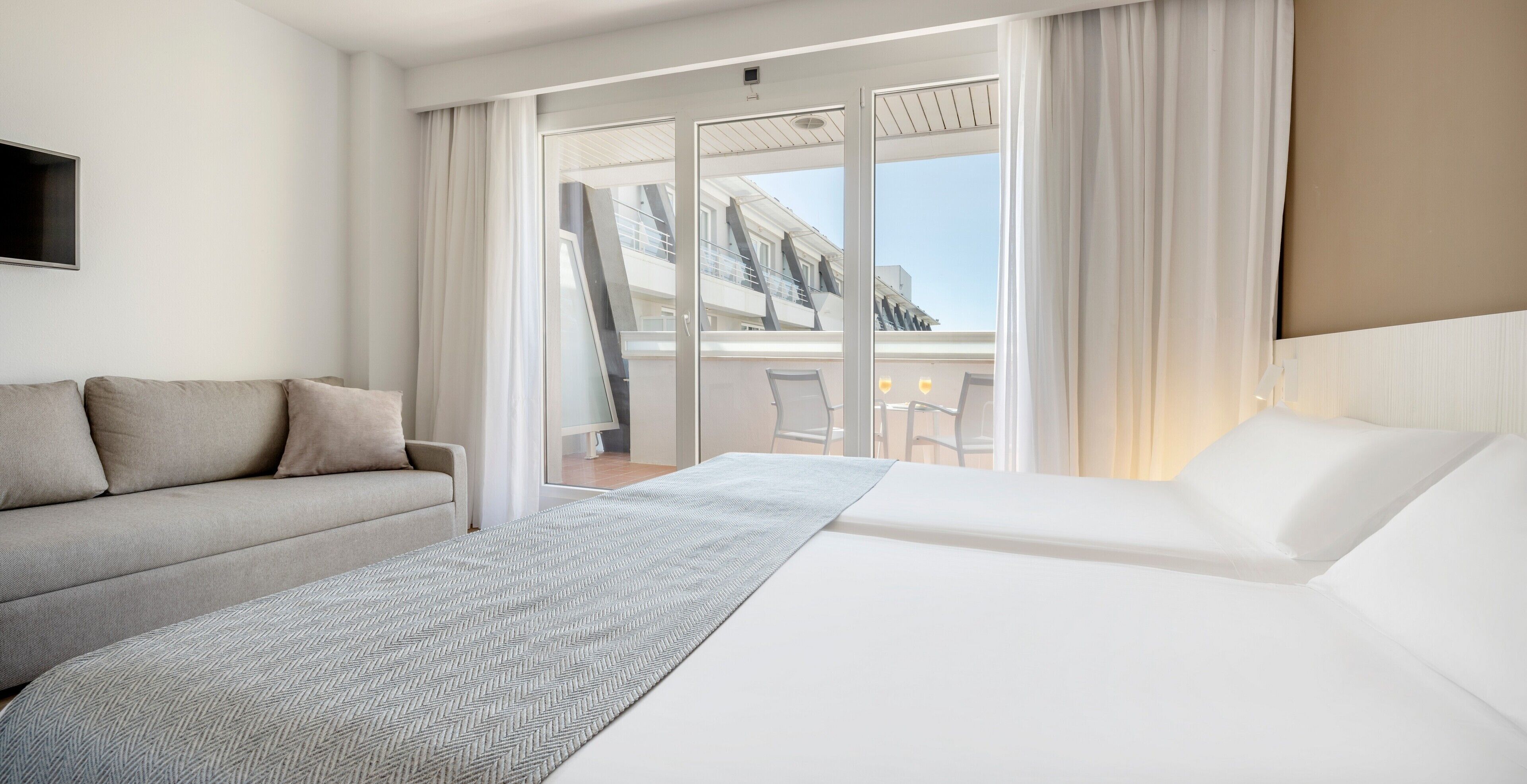 Habitación de hotel con cama, sofá, ventana grande con cortinas y vista a un edificio.
