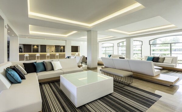 A modern hotel lobby with a central seating area, a bar, and large windows offering a view of the outdoors. - Hotel ILUNION Islantilla (Islantilla)