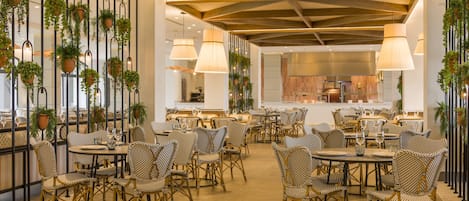 Un restaurant avec des chaises et des tables en osier, des plantes suspendues et une cuisine moderne en arrière-plan.