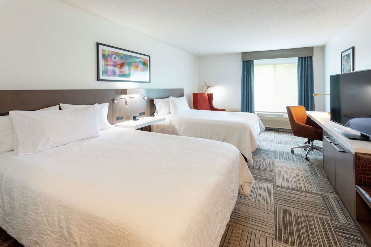 A hotel room with two beds, a desk, a TV, and a window with curtains.