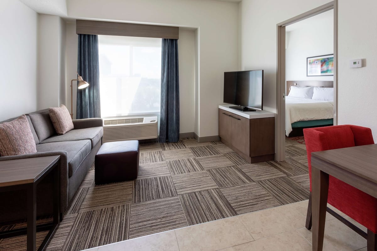 A hotel room with a sofa, a TV, a small table, a red chair, and a bed with a white duvet.
