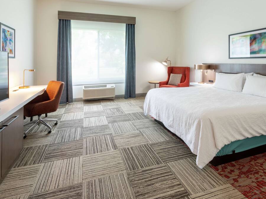 A hotel room with a bed, a desk with a TV, a chair, a lamp, and a window with curtains.
