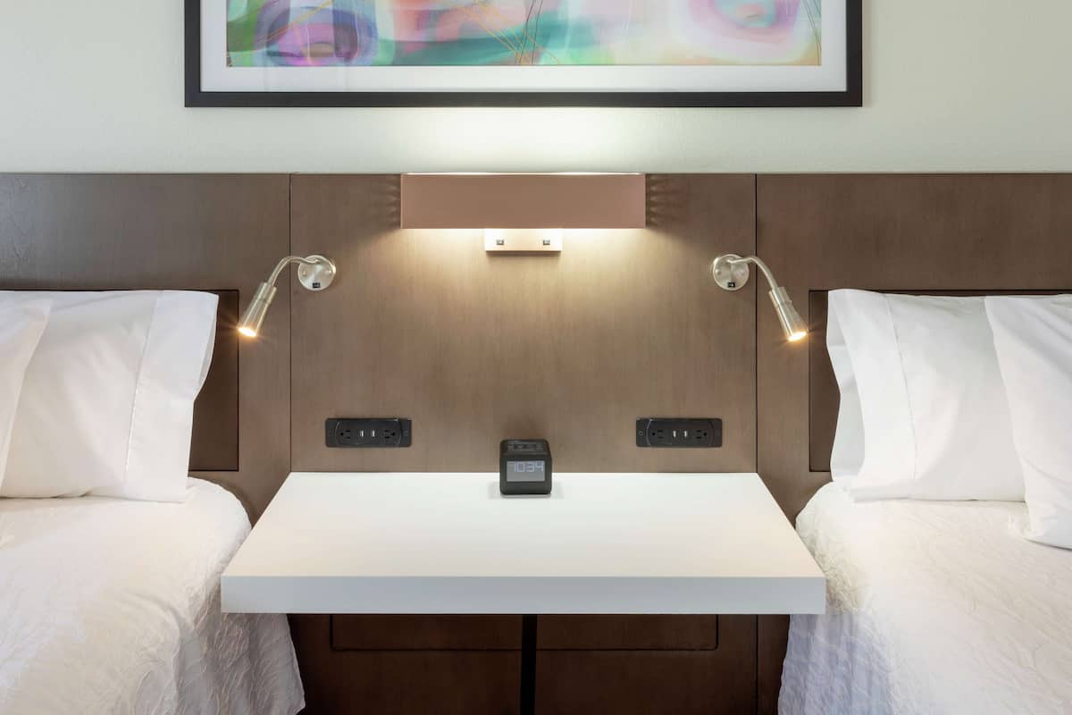 A hotel room with two beds, a bedside table, a clock, and wall-mounted lighting.