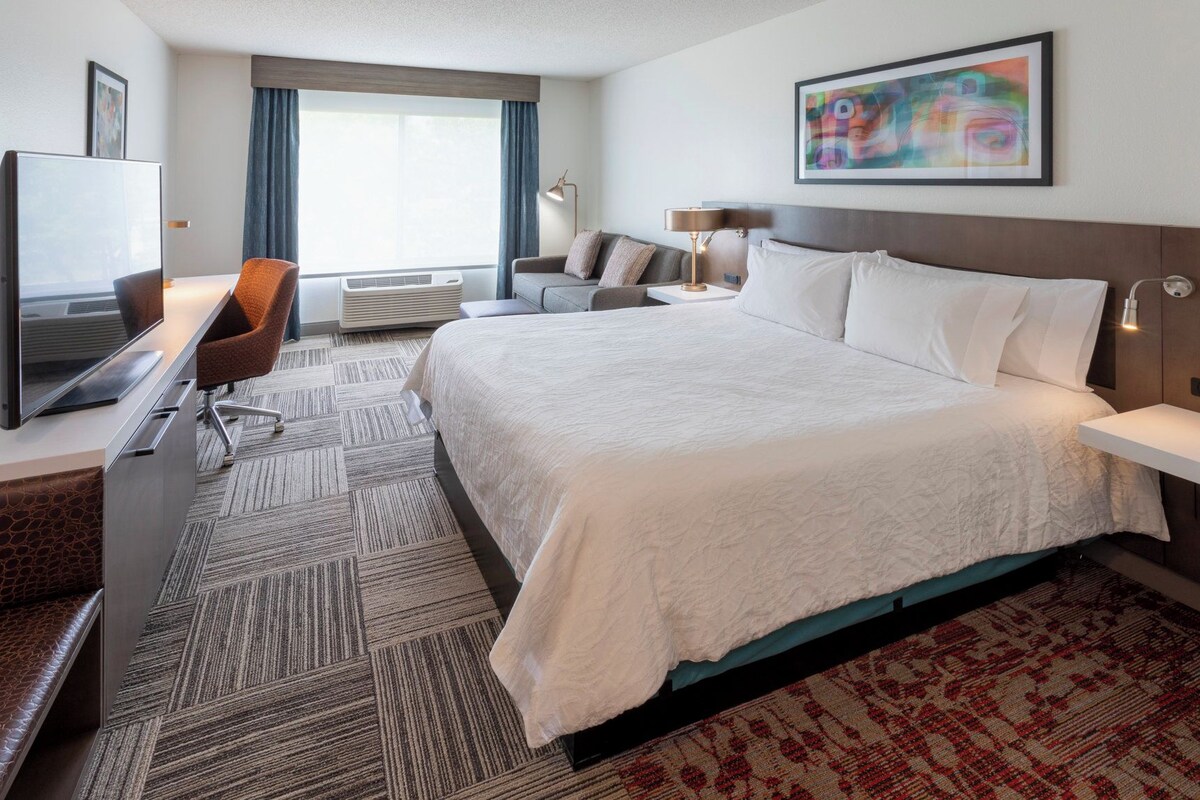 Habitación de hotel con cama, escritorio con televisor de pantalla plana, sofá y ventana grande con cortinas.