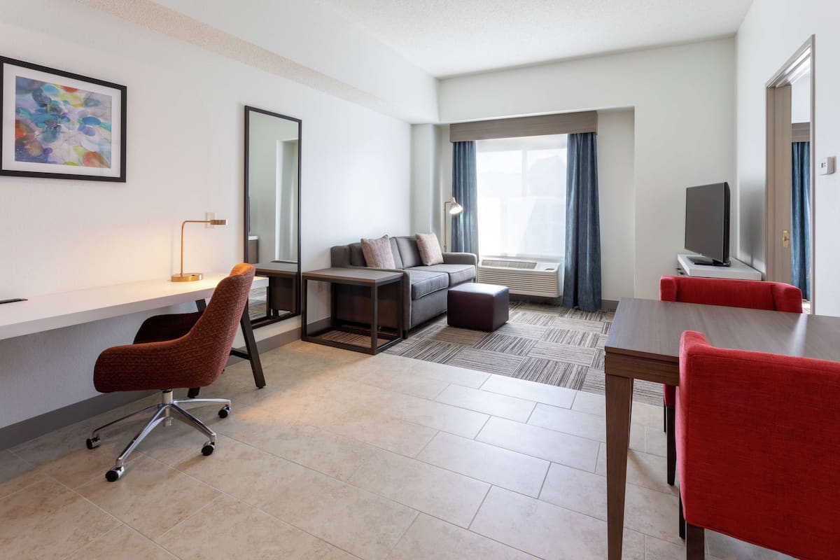 A modern hotel room with a sofa, a desk with a chair, a TV, and a large window with curtains.