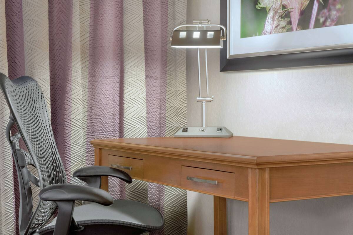 A wooden desk with a modern desk lamp, a chair, and a framed picture on the wall.
