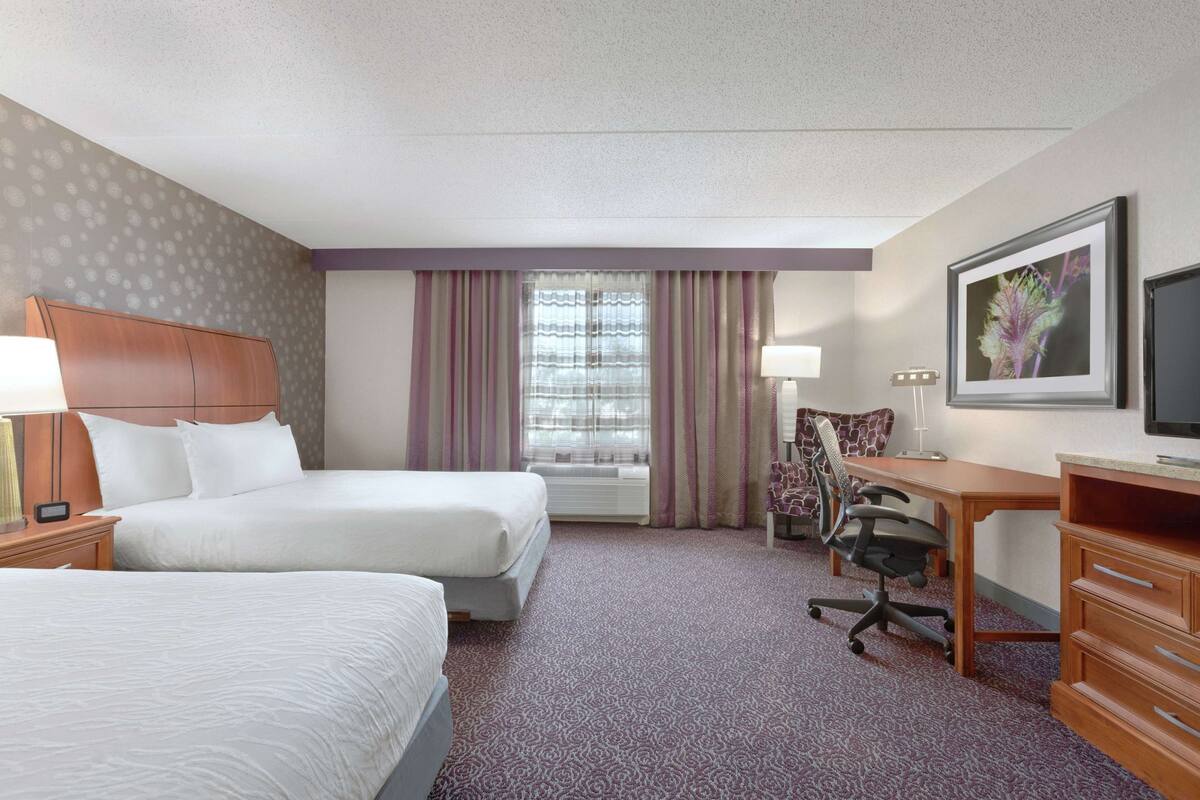 A hotel room with two beds, a desk, a chair, a TV, and a window with curtains.