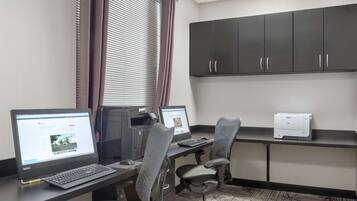 A modern office space with two desks, each with a laptop and a printer. There are two office chairs, a large black cabinet, and a window with blinds.