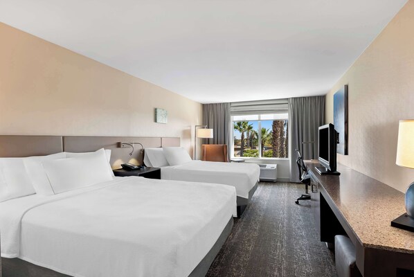 A hotel room with two beds, a desk, a TV, and a window with a view of palm trees.