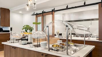 Un buffet disposé sous des présentoirs en verre transparent, avec des plats de service en acier inoxydable, et une cuisine moderne en arrière-plan.