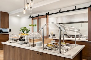 A buffet setup with clear glass covers, stainless steel serving dishes, and a modern kitchen area in the background.