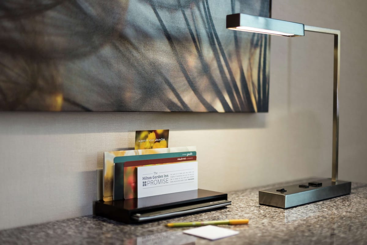 A hotel room with a modern desk lamp, a card holder with a hotel name, and a framed abstract painting on the wall.