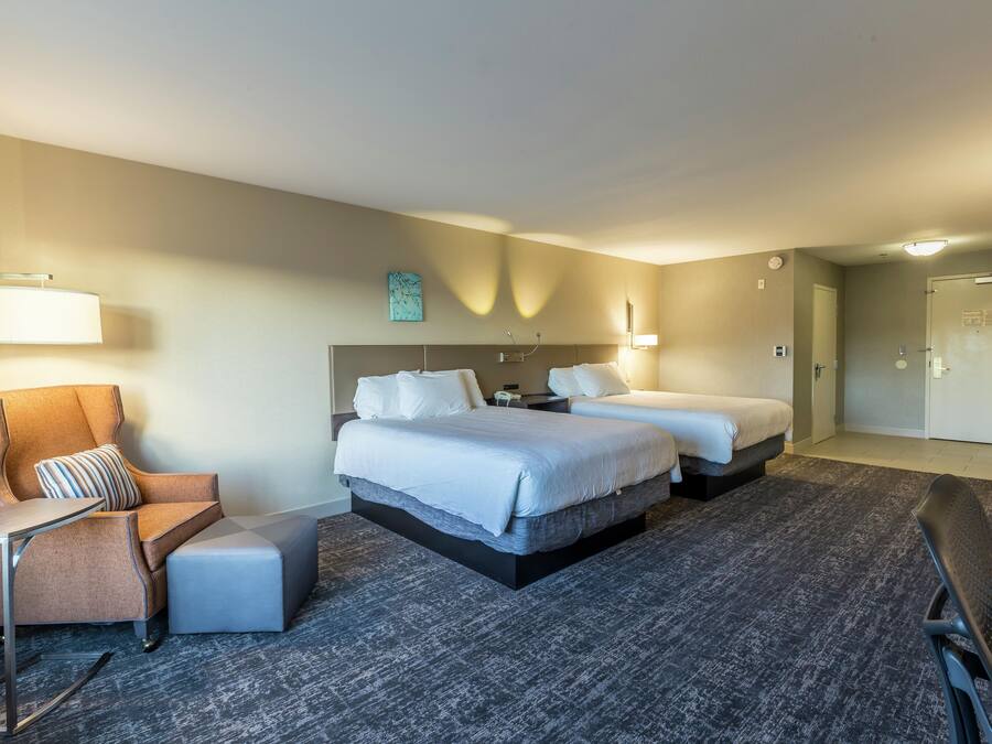 A hotel room with two beds, a chair, a desk, and a carpeted floor.