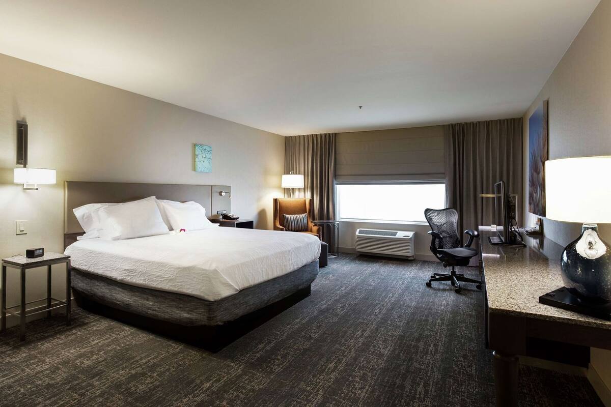 A hotel room with a large bed, a desk, a chair, a lamp, and a window with curtains.