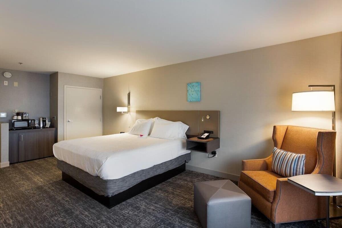 A hotel room with a large bed, a desk with a lamp, a chair, and a small table.