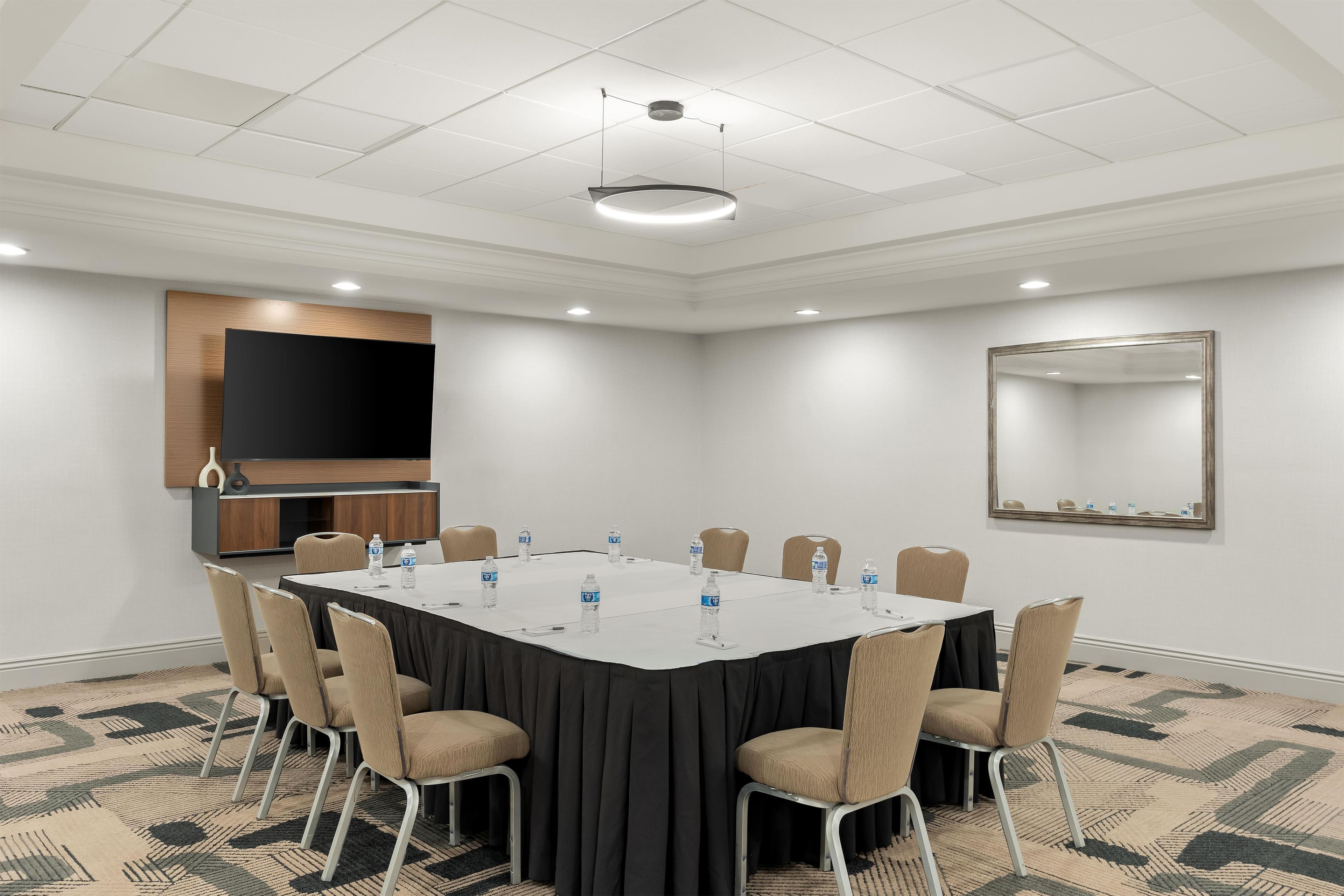 Une salle de conférence équipée d’une table ronde, de chaises, de bouteilles d’eau, d’un téléviseur à écran plat et d’un miroir au mur.