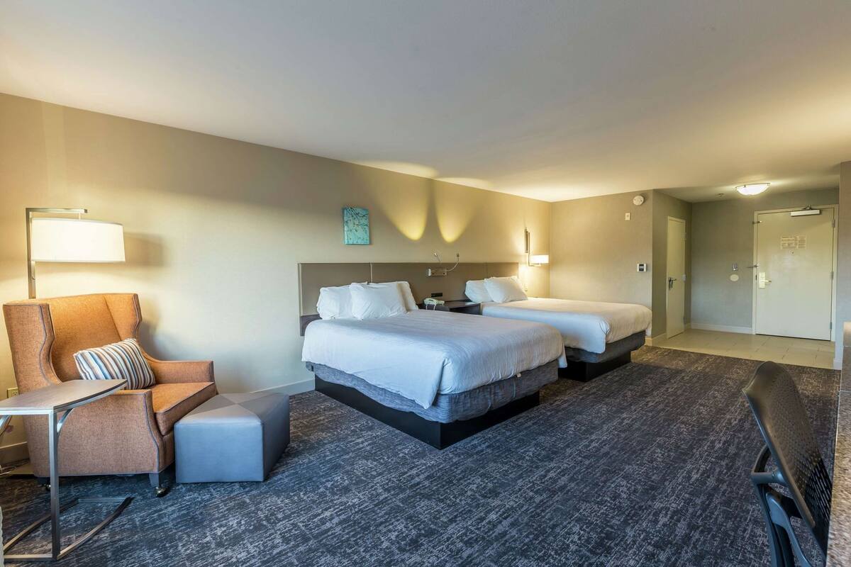 A hotel room with two beds, a chair, a desk, and a lamp.